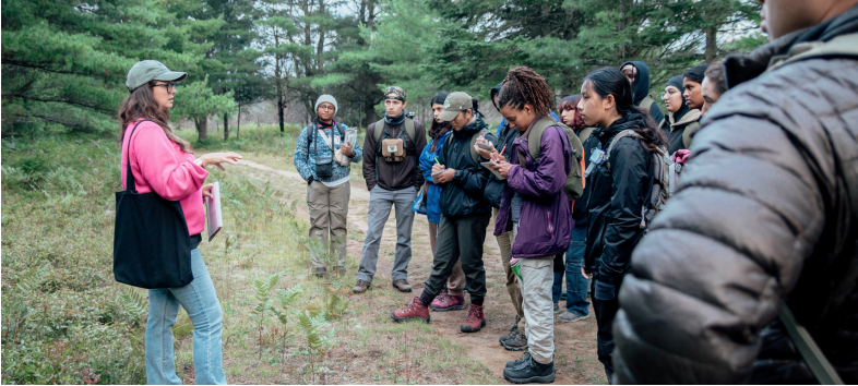 A group of people outdoors on a trail. One person is speaking to the group, while the rest of them take notes in notebooks. 